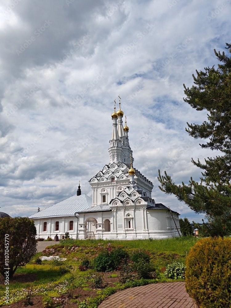 Fototapeta premium A stunning church features golden domes and intricate architecture surrounded by lush greenery under a cloudy sky