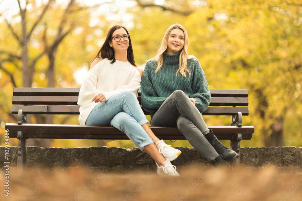 Fototapeta premium Two beautilful young women sitting on the bench in publiic park