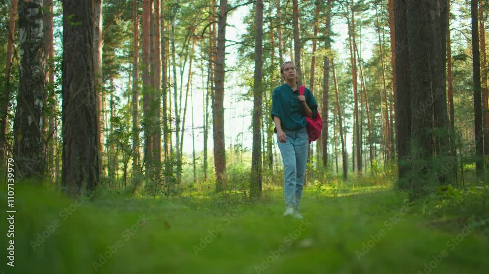 Woman strolling through forest, carrying red backpack and gazing around at tall trees bathed in sunlight, surrounded by lush greenery, she explores with a calm demeanor Woman strolling through forest, carrying red backpack and gazing around at tall trees bathed in sunlight, surrounded by lush greenery, she explores with a calm demeanor