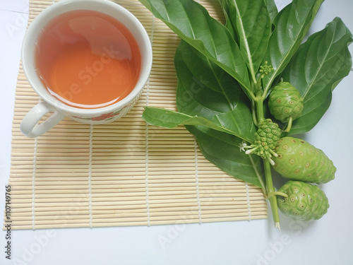 Noni fruit or Morinda citrifolia (Indian mulberry) with green leaves on tree branch on bamboo mat isolated on white background. Consuming boiled water of noni fruit juice can lower cholesterol levels photo
