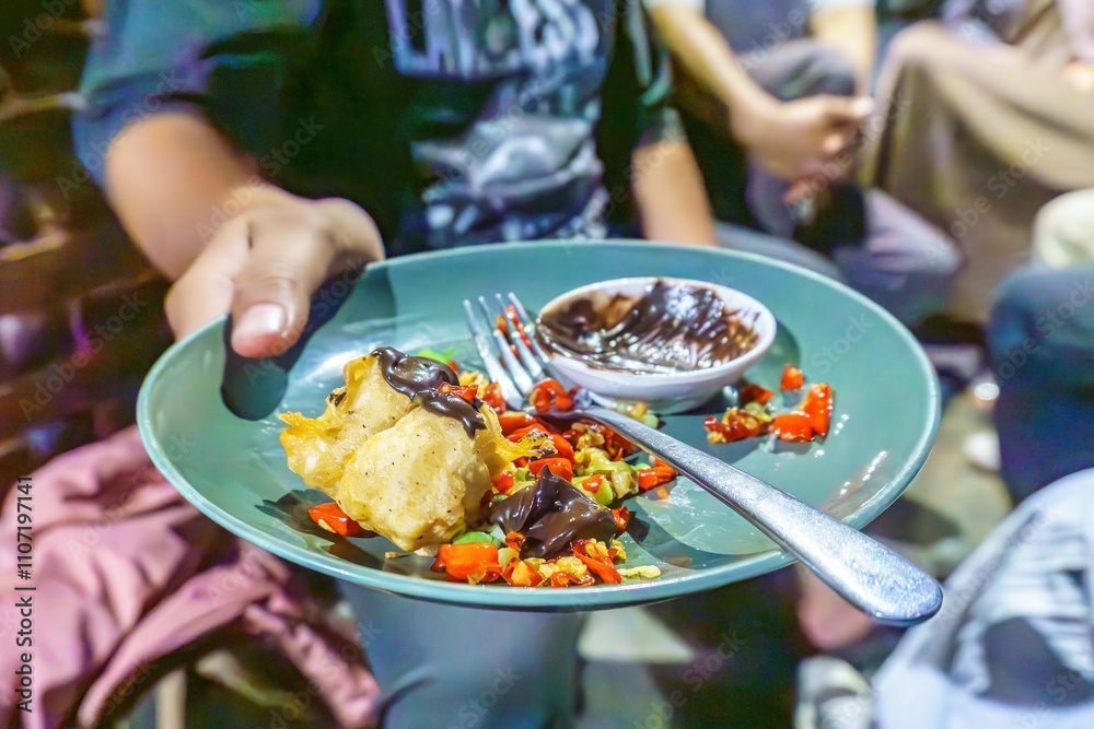 A person's hand holding a green leaf cup filled with a chocolate sauce, red chili peppers, crispy fried tofu, and a fork.