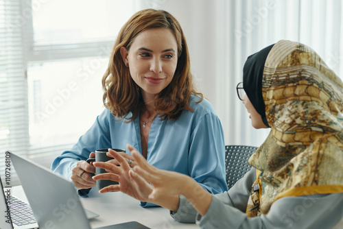 Two women engaging in discussion around laptop, expressing mutual understanding in professional environment. Natural lighting highlighting their focused interaction
