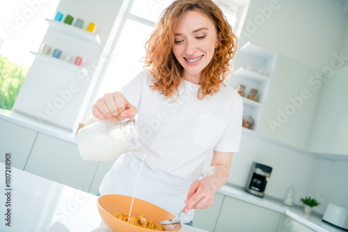 Young woman with red hair happily preparing a healthy breakfast in her bright kitchen, pouring milk into a bowl.