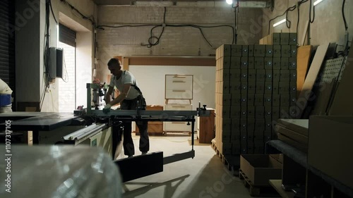 A male carpenter stands near a machine in a furniture factory workshop and cuts wooden elements with the machine. Conveyor production, craft, small business. carpentry workshop, industrial equipment, 