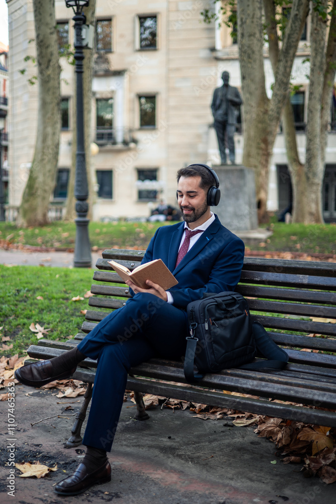 Obraz premium Businessman wearing headphones reading book while sitting on bench in park