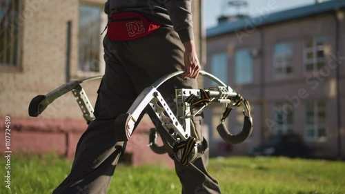 A Guy carrying large metallic spring stilts, wearing black pants, dark gray long-sleeved shirt, and red hip bag with visible white logo. Background is blurred with grassy area and distant building photo