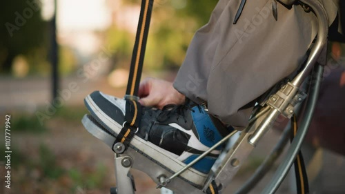 Close-up of person fastening shoes to spring stilts using orange and black straps, wearing gray trousers and long-sleeved shirt outdoors, blurred background, focus on adjustment process photo