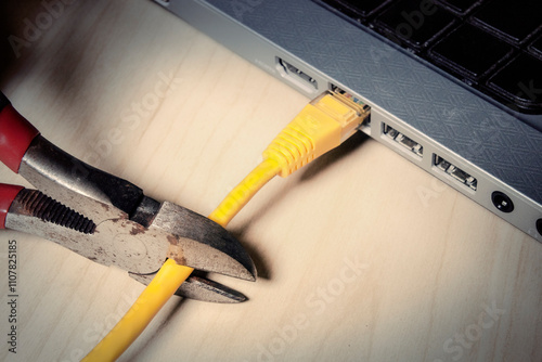 Sharp pliers severing yellow ethernet cable connected to laptop, dramatic angle. Stark composition symbolizing digital disconnection, censorship and network interruption.

