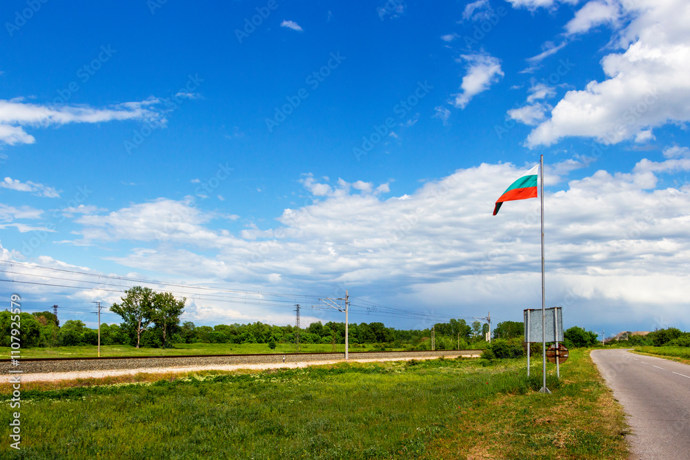 Fototapeta premium Railway tracks and the Bulgarian national flag under cloudy May sky at the exit of the village of Krum in direction of the town of Dimitrovgrad in Haskovo Province, Southern Bulgaria