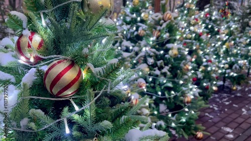 video postcard with festive Christmas tree decorated with striped gold and silver balls with sparkling garlands near the wall of the house against the background of decorated small trees on the path 