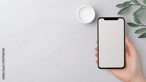 Hand holding a black smartphone with a blank white screen. the phone is placed on a white marble surface with a small white bowl and a green plant with leaves on the right side.