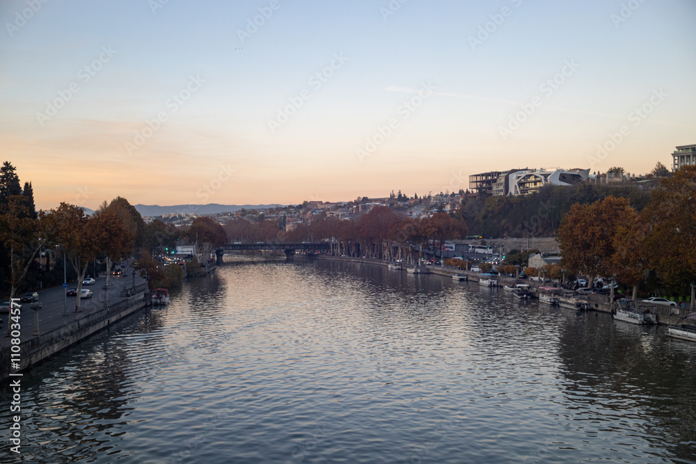 Obraz premium Serene autumn twilight envelops Tbilisi as golden leaves float by the river, painting a peaceful urban landscape.