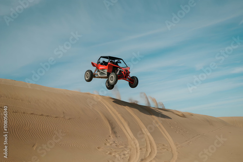 Fototapeta Naklejka Na Ścianę i Meble -  Red ATV mid air, leaping off golden dunes under a clear blue sky