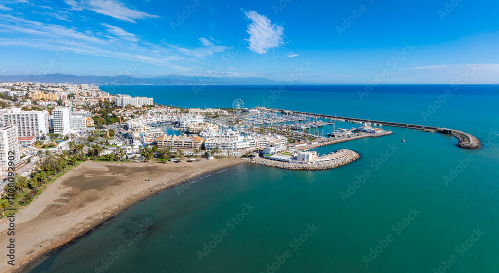 Fototapeta premium The image captures Benalmadena's marina with boats, sandy beaches, and white Mediterranean buildings. A Ferris wheel stands near the vibrant blue sea.