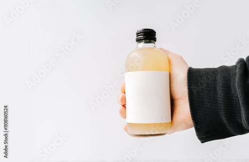 Woman hand holding small transparent glass bottle with black cap and white label, filled kombucha of yellow