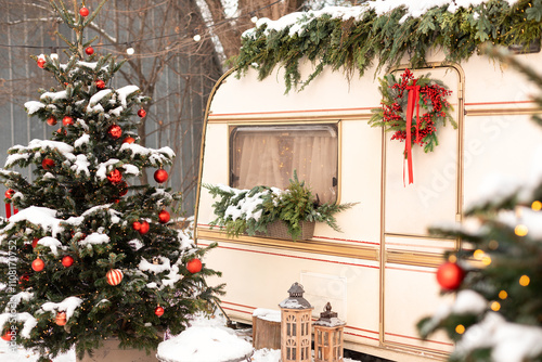 Xmas wreath on door. Exterior design travel trailer or recreational vehicle with Christmas decorations, decorated Xmas tree with balls in pot in snow yard. Cozy festive mobile rv home camping winter.