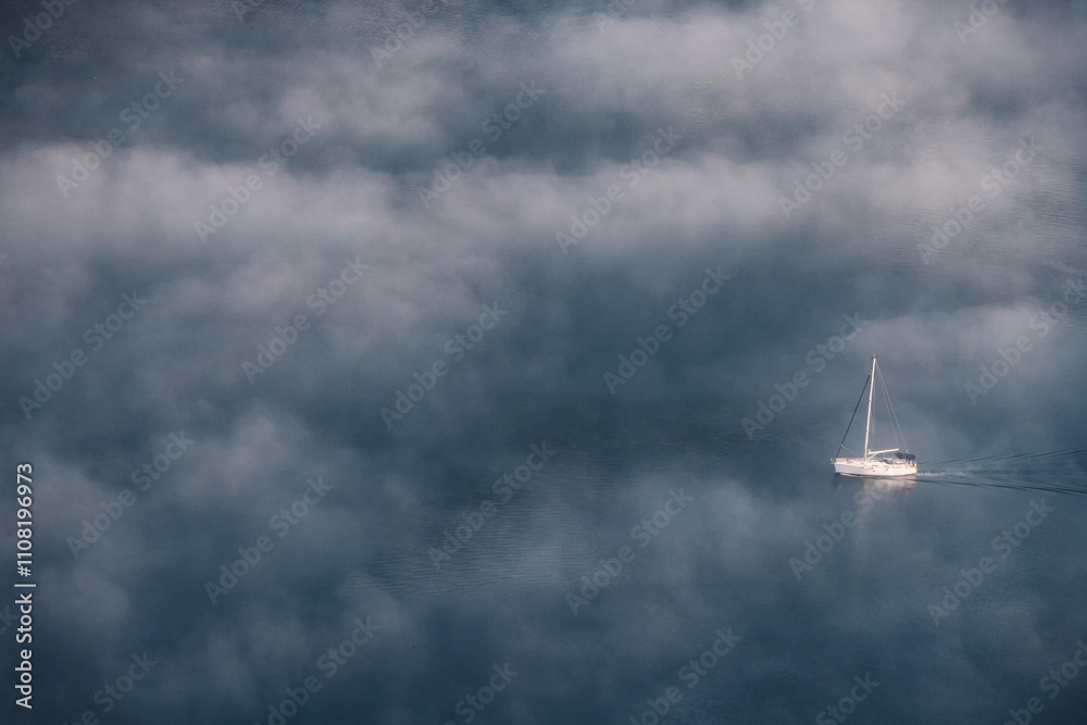 White sailboat sailing on calm sea with low clouds creating a mystical atmosphere