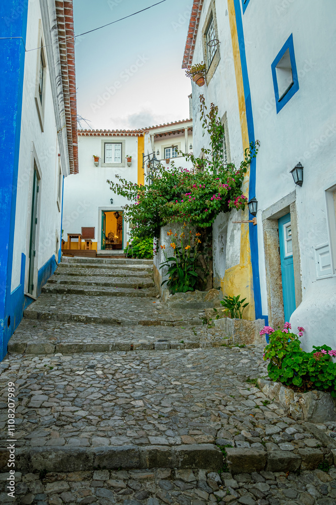 Fototapeta premium Cobblestone alley climbs between colorful houses adorned with vibrant bougainvillea, creating a charming scene in the medieval town of Obidos, portugal