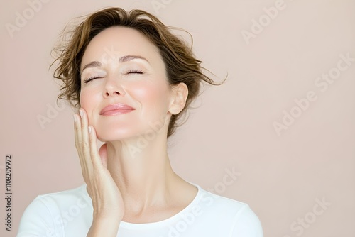 Woman 45 years old face care. She is gently touching her face, expressing relaxation and contentment. The simple backdrop enhances her calm demeanor, reflecting a moment of self-care. photo
