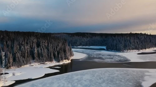 An aerial winter view of a forest landscape with a frozen river bordered by snow-laden pines. 