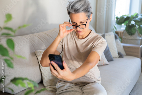 A middle-aged woman sits on the couch and lifts her glasses trying to read a message on the phone.