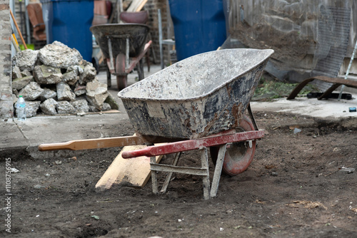 A worn wheelbarrow sits on a busy construction site filled with debris and rock piles, conveying hard work and industry.