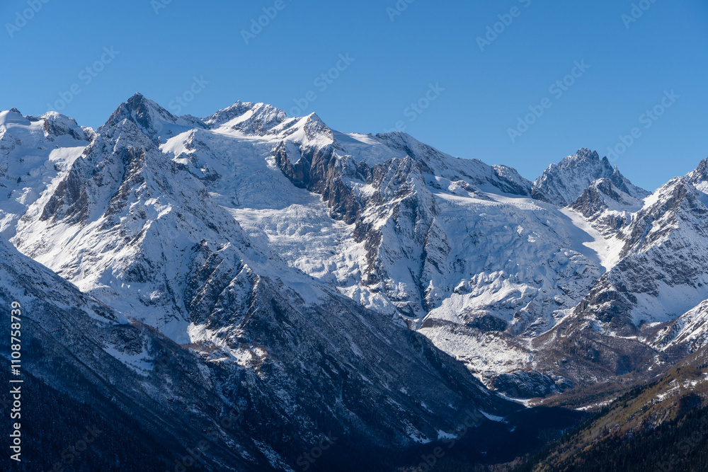Fototapeta premium Snowy peaks and glaciers cover giant rocks. Pine and spruce forests grow at foot of mountains. which Majestic mountains against blue sky. Sunny day. Autumn 2024. Karachay-Cherkessia. Dombay.
