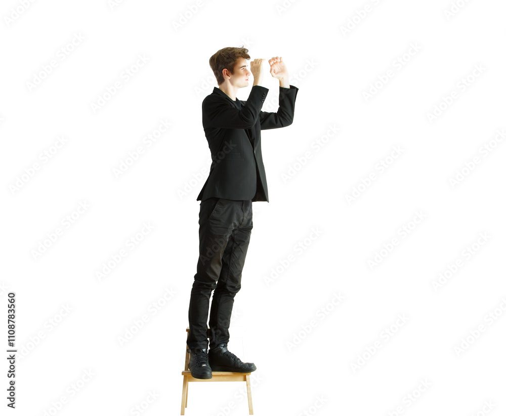 Obraz premium Full-length image of a man in asuit standing on a wooden chair, reaching upward, against a white background. Concept of effort or problem-solving