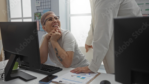 Woman smiling at colleague in modern office while discussing work, with documents and computers indicating a collaborative business environment