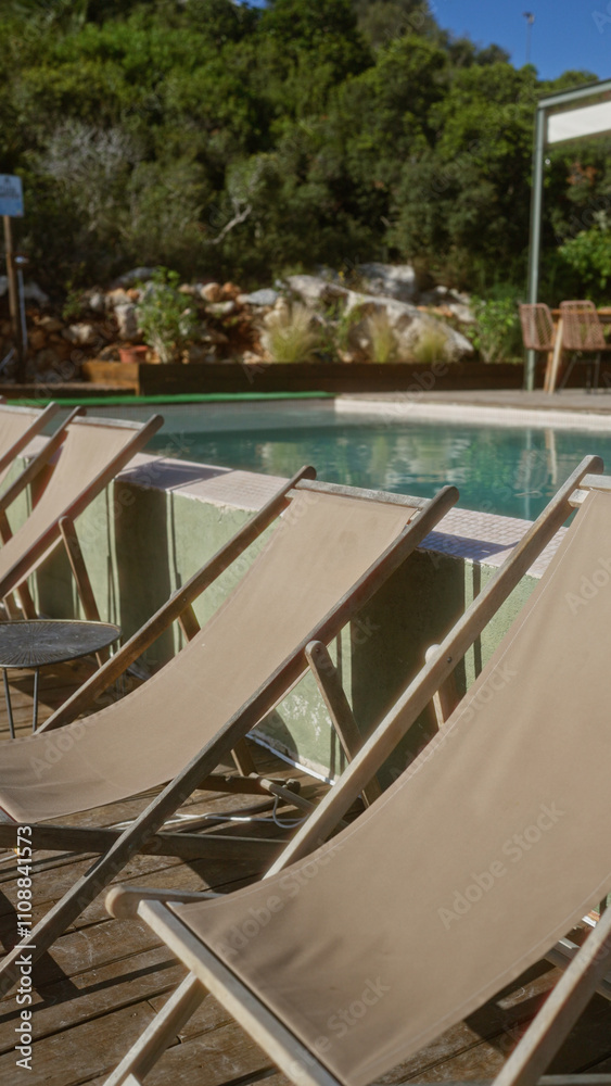 Empty deck chairs by an outdoor swimming pool set against lush greenery and sunny skies, perfect for relaxation and leisure in a serene natural setting. Empty deck chairs by an outdoor swimming pool set against lush greenery and sunny skies, perfect for relaxation and leisure in a serene natural setting.