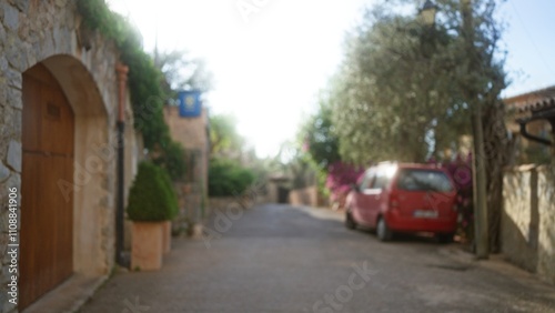 Blurred street scene in mallorca, featuring a defocused red car and stone buildings under a vibrant blue sky, with lush greenery and bokeh effects creating a serene village atmosphere.
