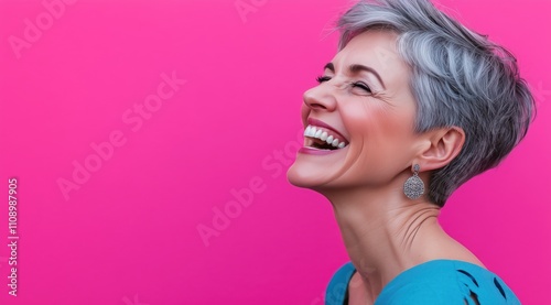 Femme d'une cinquantaine d'années riant, aux cheveux courts gris, portant un vêtement bleu et des boucles d'oreilles sur un fond rose. photo