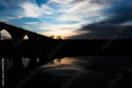 Silhouette of an Arched Bridge at Sunset Reflecting on Calm Waters in Fuentes Claras, Avila, CyL