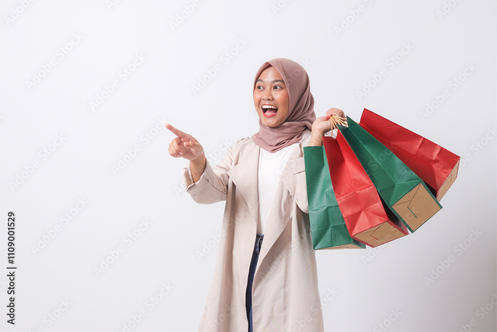 Portrait of excited Asian hijab woman in casual suit holding paper shopping bags while pointing with finger. Shopaholic girl and discount buying concept. Isolated image on white background Portrait of excited Asian hijab woman in casual suit holding paper shopping bags while pointing with finger. Shopaholic girl and discount buying concept. Isolated image on white background