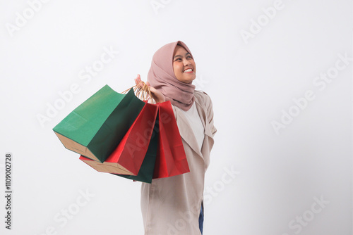 Portrait of excited Asian hijab woman in casual suit holding and showing colorful paper shopping bags. Shopaholic girl and discount buying concept. Isolated image on white background
