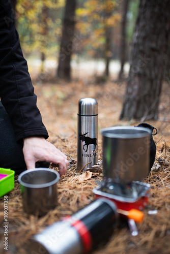 A man prepares a hot drink on a gas stove in a metal cup. A metal thermos. In the forest.
