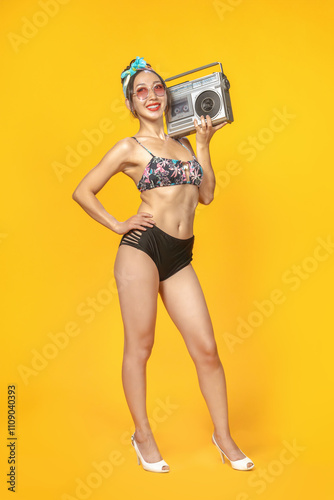 Hot Asian girl in a swimsuit and with a headband with a retro tape recorder on a bright yellow background. Isolated