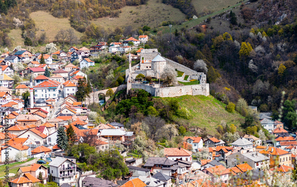 Fototapeta premium Scenic view of Travnik, Bosnia and Herzegovina, featuring historical buildings, mosques with minarets, a clock tower, and a fortress surrounded by lush green hills. A picturesque Balkan town.