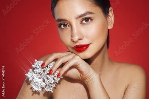 Beautiful young woman with red lipstick, shiny clean skin and face, showing glowing snowflake, near face and smiling, isolated on a red background. Christmas holidays concept