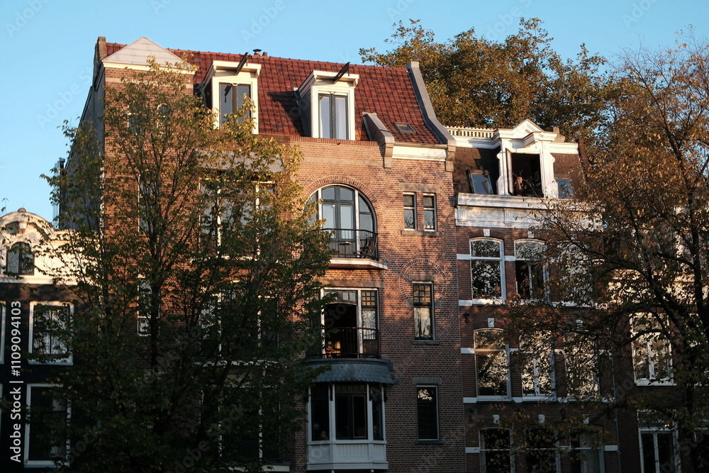 Fototapeta premium Fassaden schöner alter Wohnhäuser mit Mansarde, Dachgaube und alten Bäumen im Herbst im Licht der Abendsonne an der Keizersgracht in der Altstadt von Amsterdam, der Hauptstadt der Niederlande