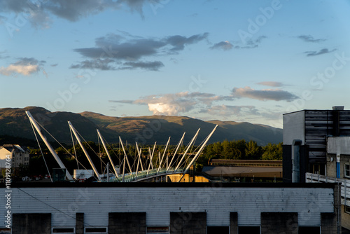 This wide angle photograph offers a sweeping view of a footbridge in front of a mountain backdrop against a cloudy sky and a building in the foreground.