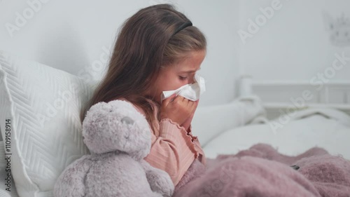 Sick girl sneezing into tissue in bedroom at home. Female child blowing her runny nose, suffering from cold and flu, while using a digital tablet