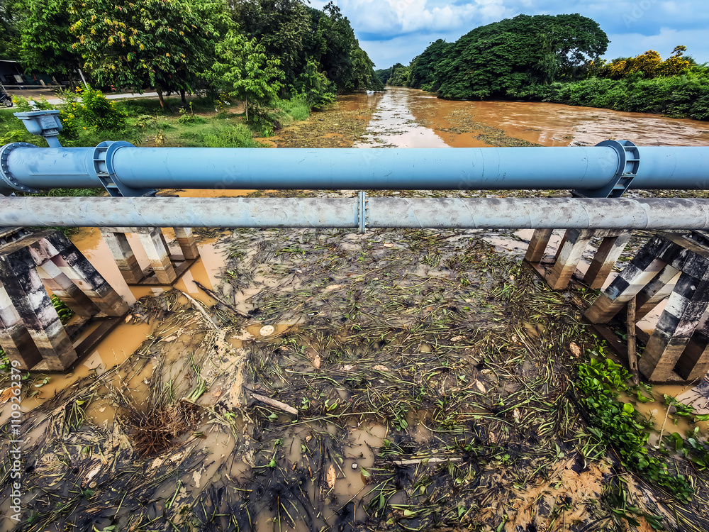 View of irrigation canal with weeds and debris flowing together with strong flowing dirty brown water due to soil and mud from forest after heavy rains and floods. La Nina crisis, Water pollution. View of irrigation canal with weeds and debris flowing together with strong flowing dirty brown water due to soil and mud from forest after heavy rains and floods. La Nina crisis, Water pollution.