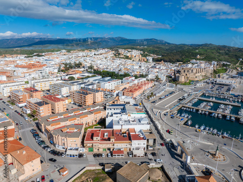 Aerial perspective of Tarifa, Spain, highlighting Guzman Castle, a marina with boats, whitewashed buildings, rolling hills, and wind turbines. photo