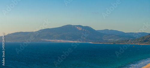 Aerial view of Tarifa, Spain, featuring the ocean and Sierra de San Bartolome. Kite surfers dot the clear blue waters, adding movement to the scene.