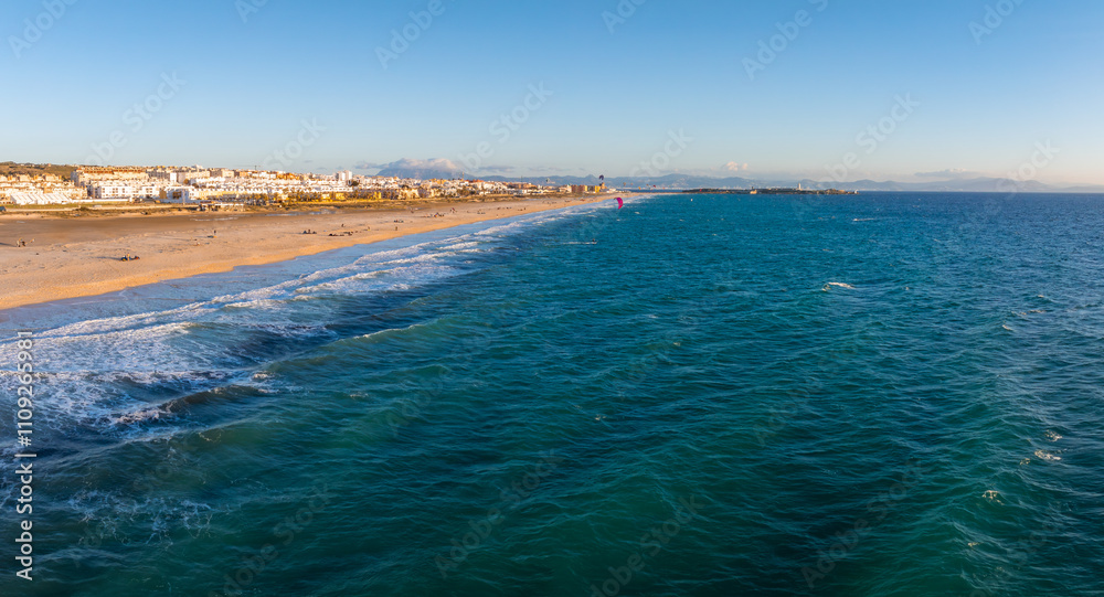 Tarifa's sandy beach stretches along the coast, with kite surfers on turquoise waters. White buildings and distant mountains are visible under a clear blue sky. Tarifa's sandy beach stretches along the coast, with kite surfers on turquoise waters. White buildings and distant mountains are visible under a clear blue sky.