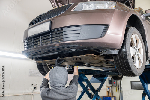 Mechanic in hoodie and gloves working on a car's undercarriage in a repair shop. Car is lifted on a platform. Concept of car repair photo