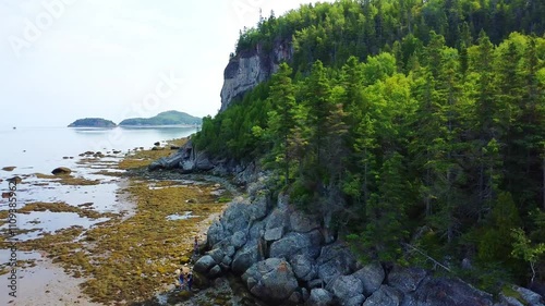 A family of tourists walks on a rugged rocky beach filled with seaweed only walkable at low tide in a panoramic coastal setting.