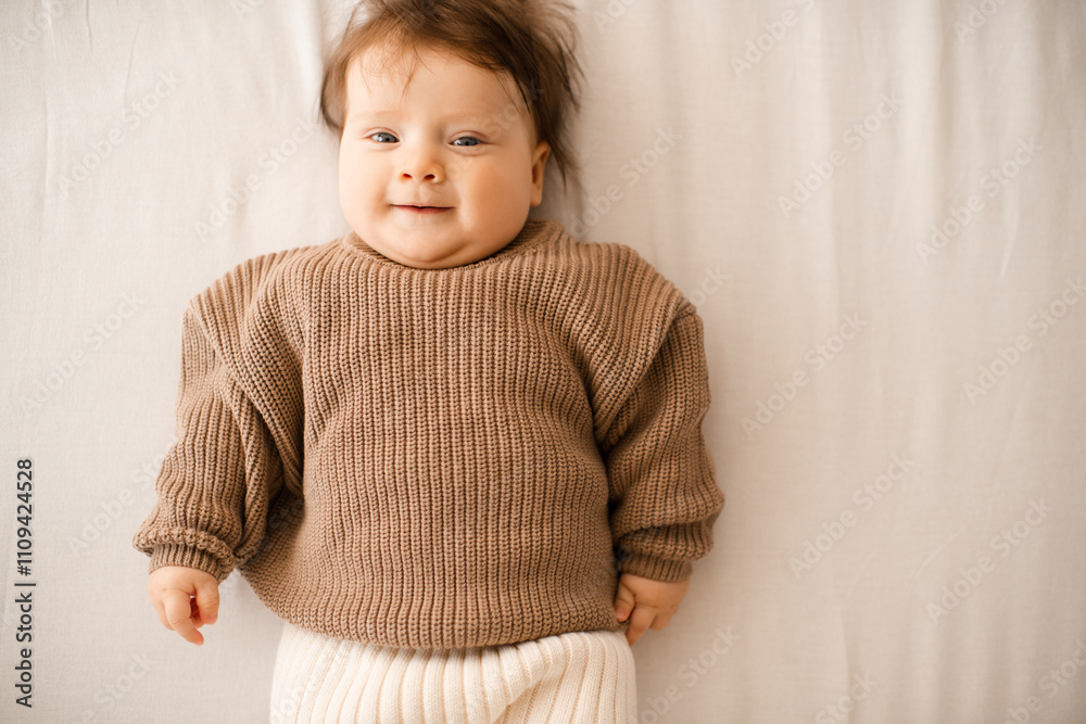 Fototapeta premium Smiling baby girl 5-6 months old lying in bed on white blanket wearing knitted sweater close up. Childhood.