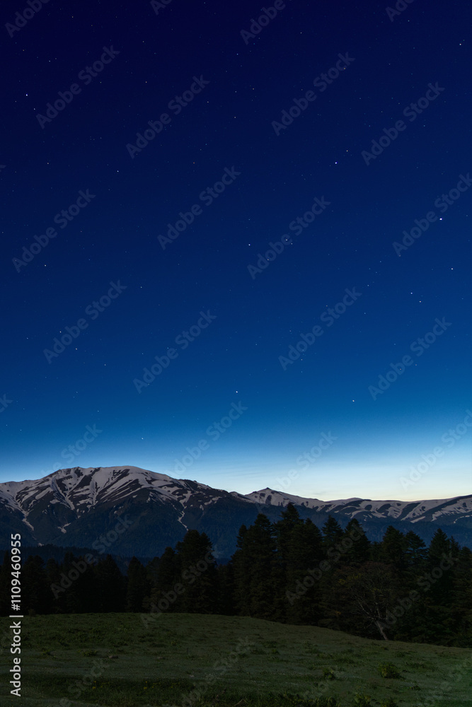 A serene night sky filled with stars above a mountainous landscape of Zekari Pass. The mountains have snow-capped peaks, and there is a forested area at the base of the mountains. A serene night sky filled with stars above a mountainous landscape of Zekari Pass. The mountains have snow-capped peaks, and there is a forested area at the base of the mountains.